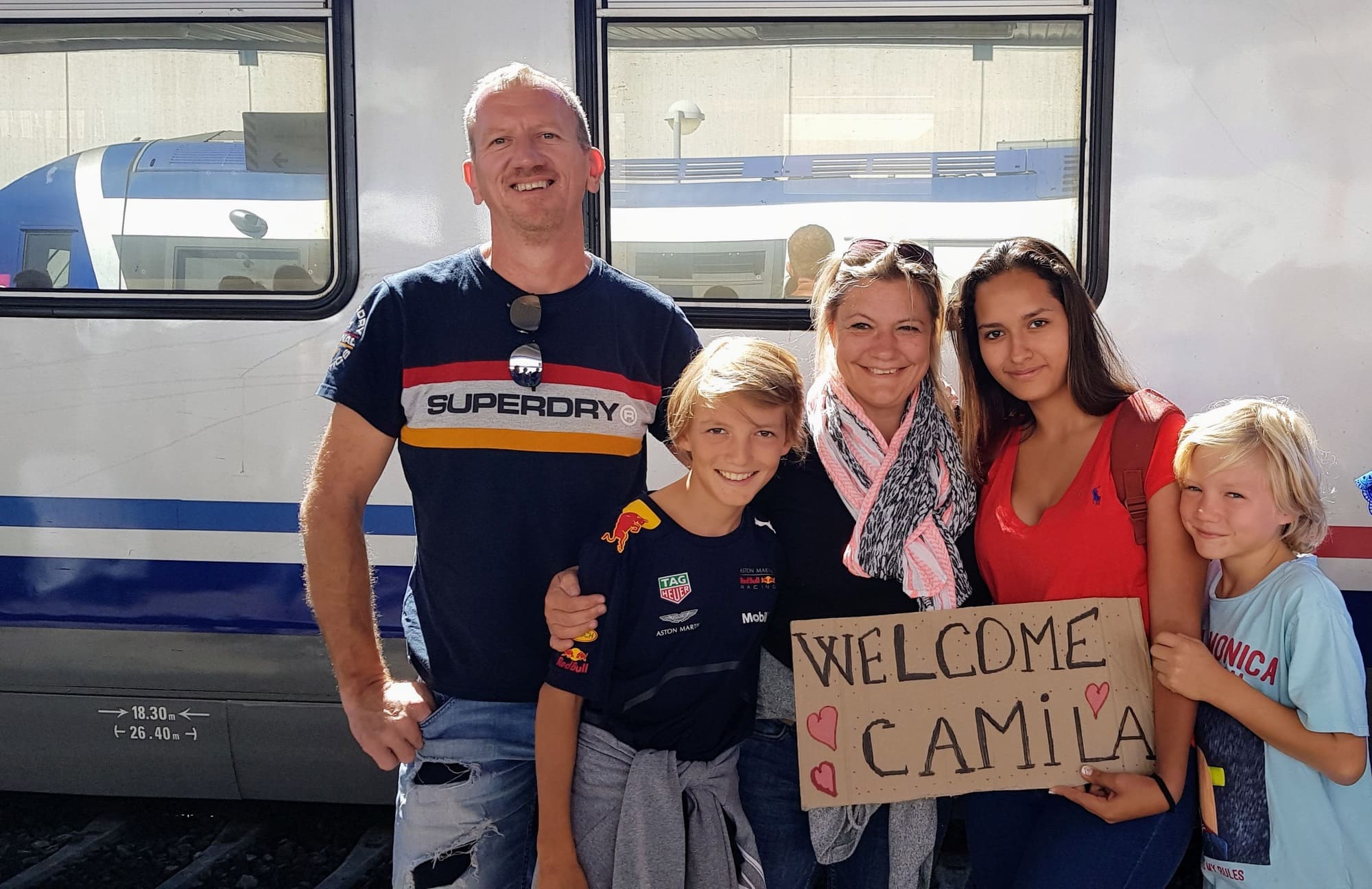 Famille française souriante accueillant Camila, une jeune Bolivienne, à la gare avec une pancarte “Welcome” — symbole d’un accueil chaleureux et de liens sans frontières