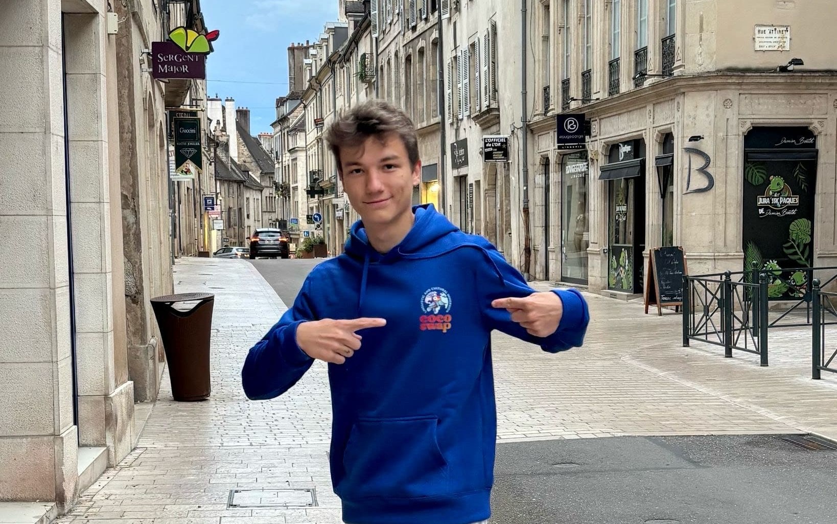 Loup, parti aux Pays-Bas avec Coco Swap, sourit dans une rue européenne en portant le sweat bleu de la plateforme – symbole de respect et d’engagement entre utilisateurs