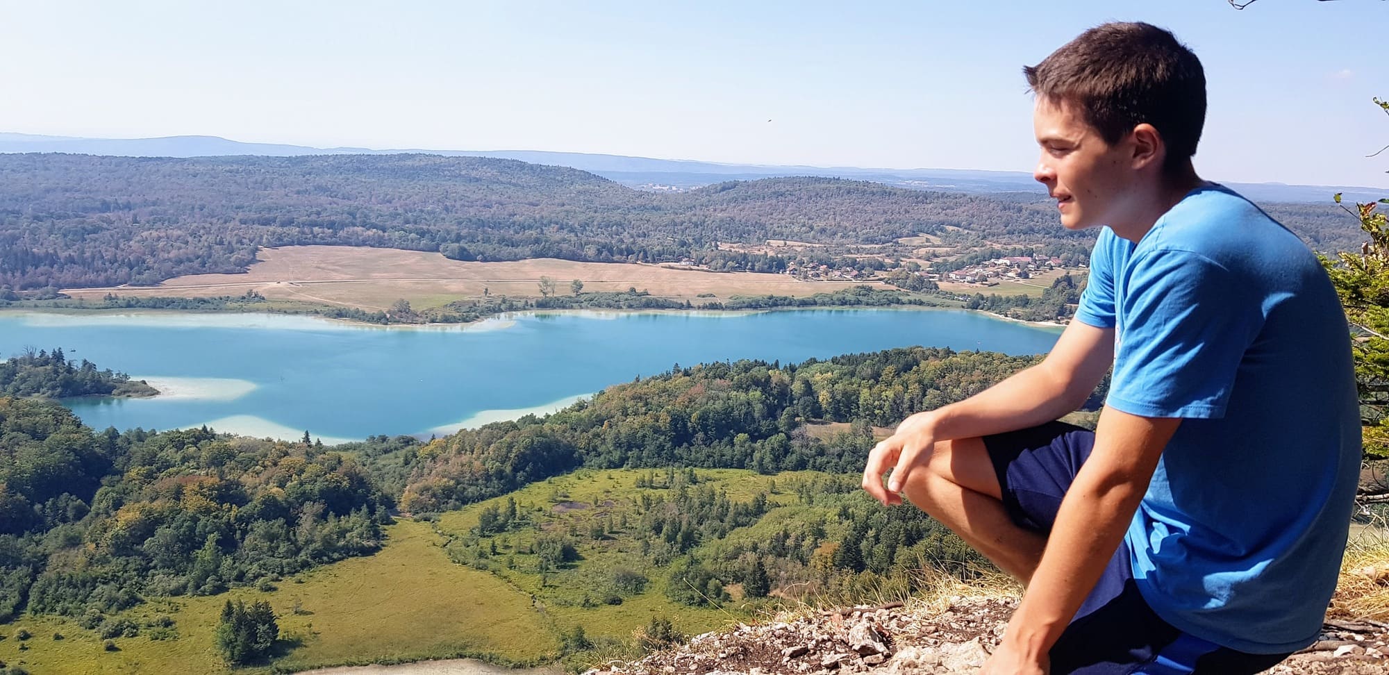 José, jeune Espagnol accueilli en France, contemple un lac pendant son séjour chez Elio — une amitié née d’un échange réciproque entre familles françaises et espagnoles